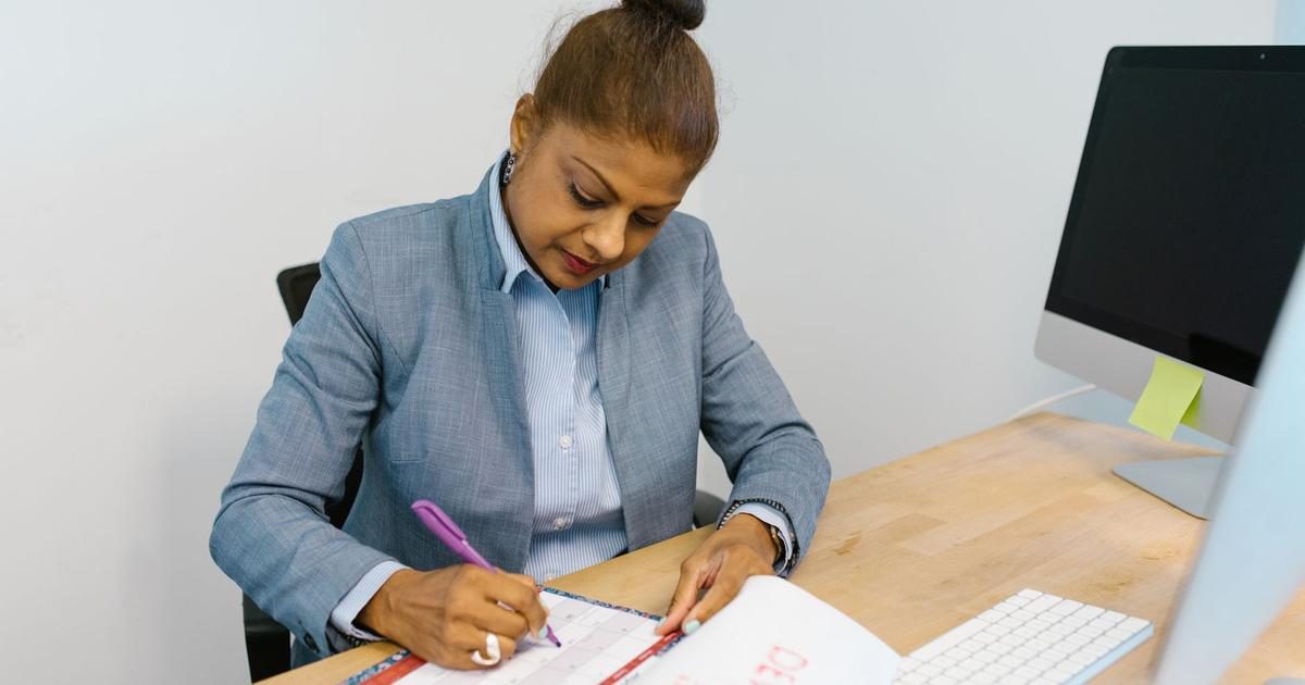 An office manager in a gray blazer marks dates on a paper calendar at her desk beside a desktop computer.