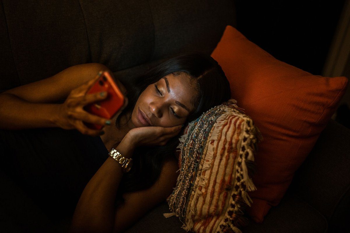 A woman lies on a couch late in the evening, lit by her phone screen as she scrolls through it.
