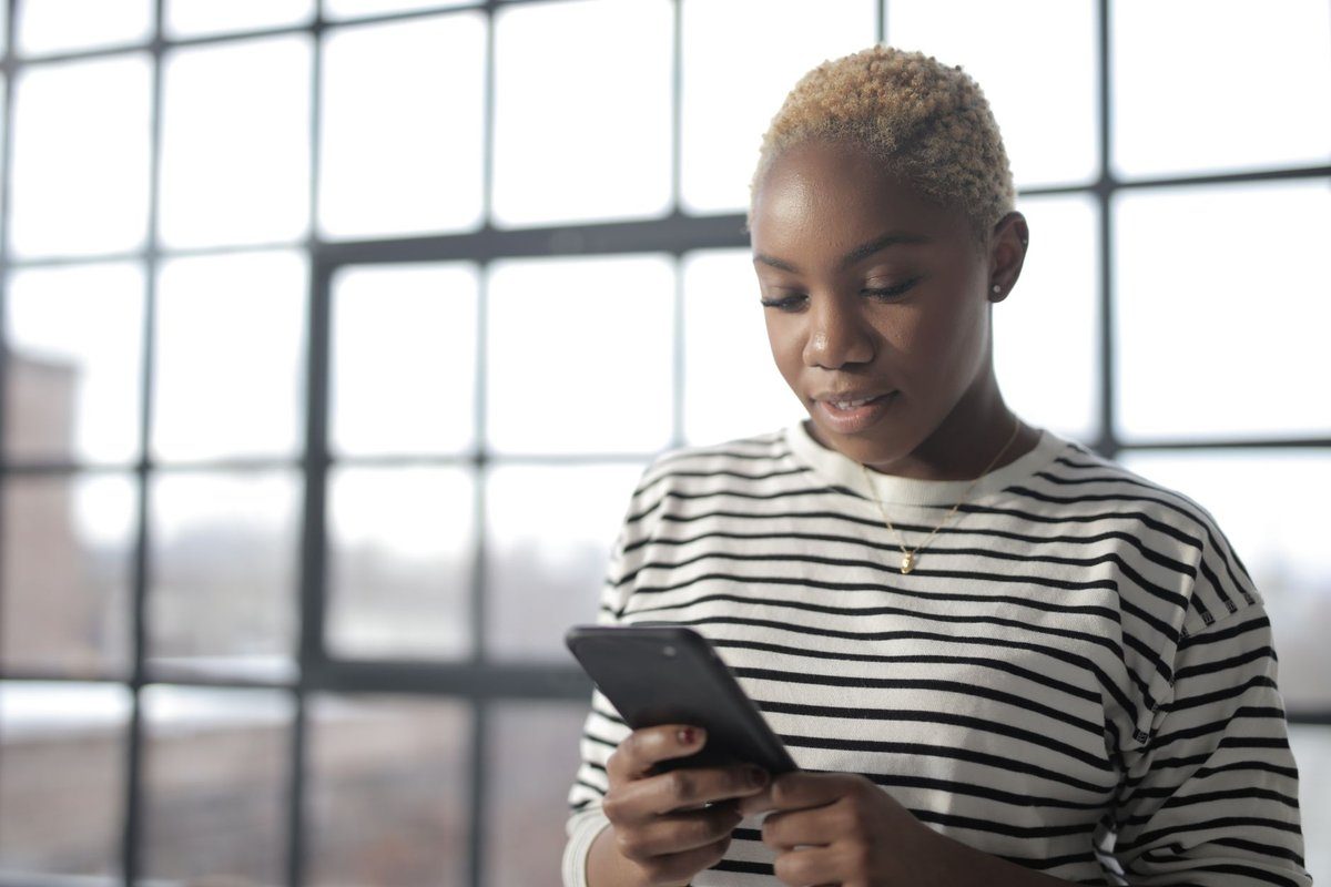 A caregiver in a striped shirt reads a message on her smartphone beside a large window, deciding whether to accept an open shift.