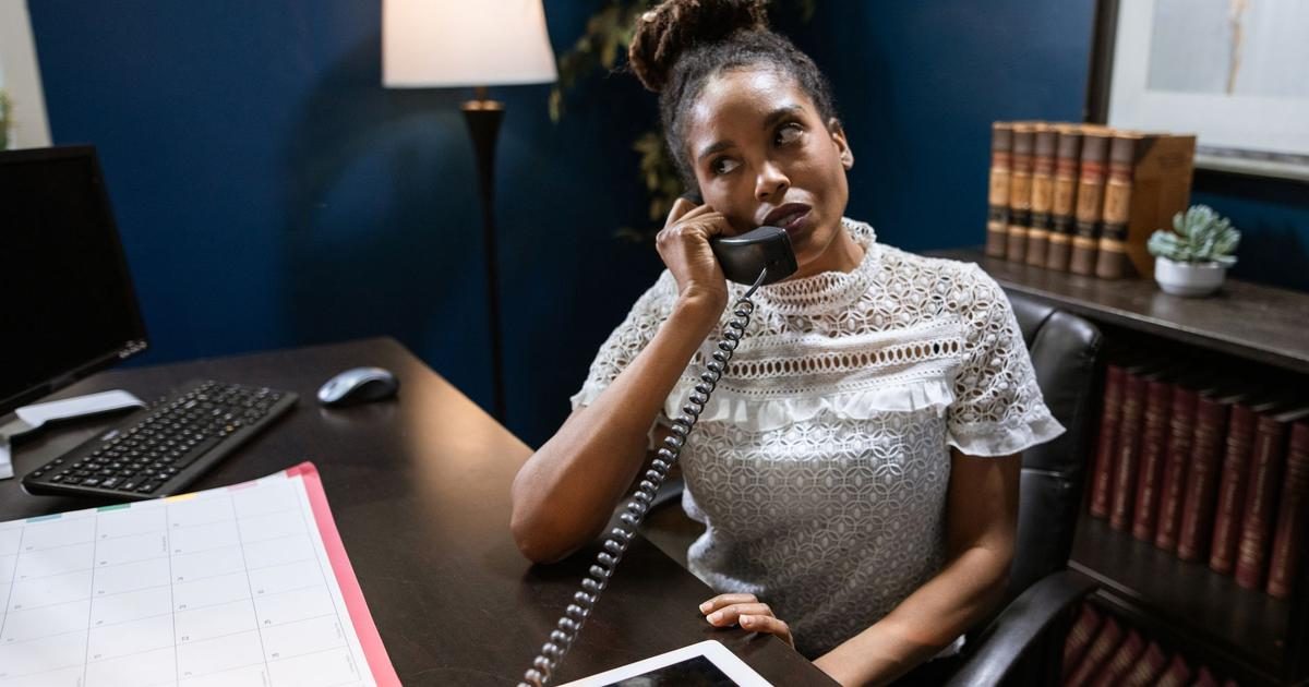 A home care scheduler holds a desk phone to her ear with a monthly planner open in front of her as she works out who can cover a shift.