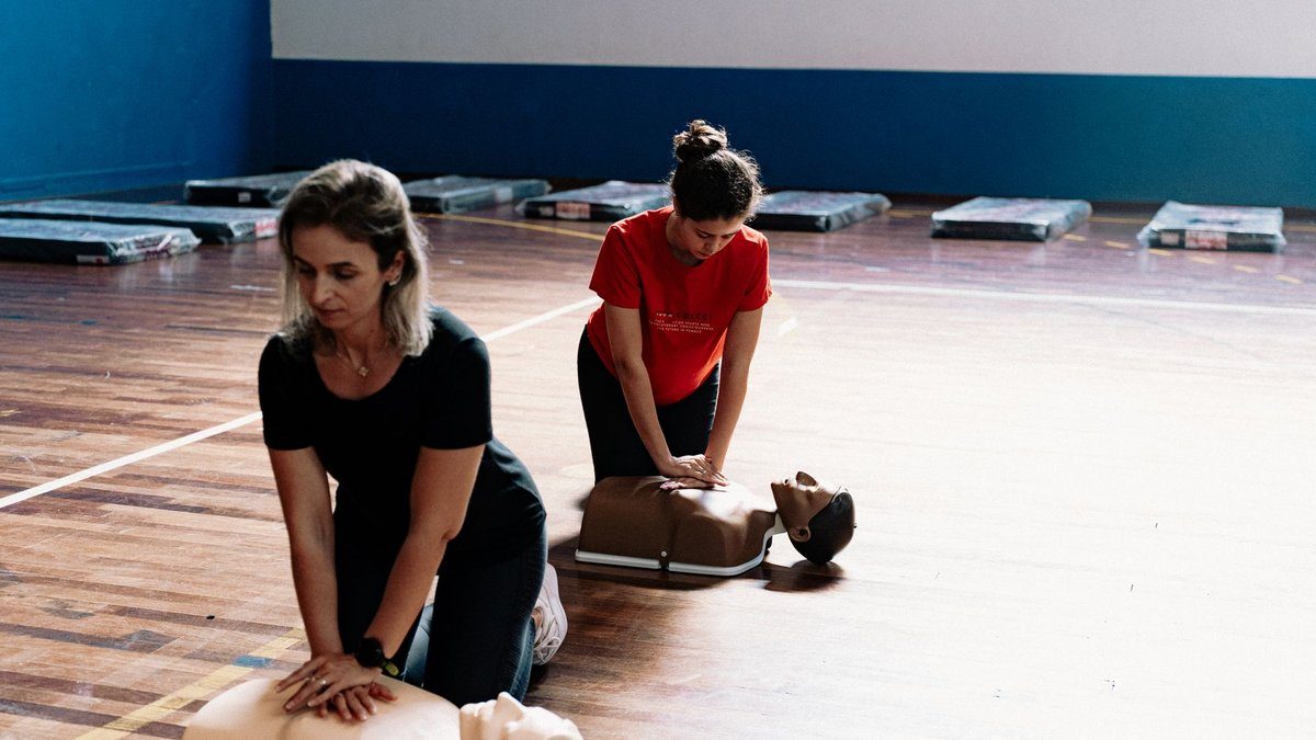 Two women kneel on a gym floor practicing chest compressions on CPR training mannequins during a first aid class.