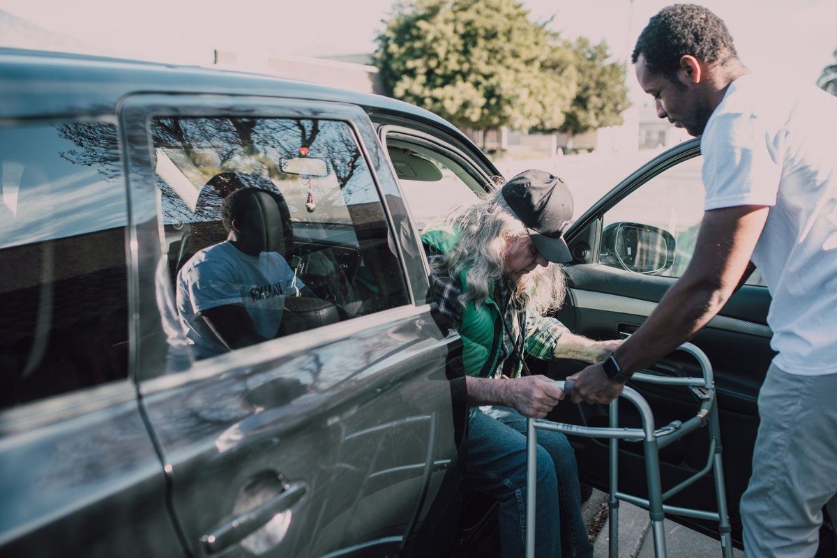 A caregiver in a white shirt steadies a walker as an older man in a cap climbs out of the passenger seat of a car.