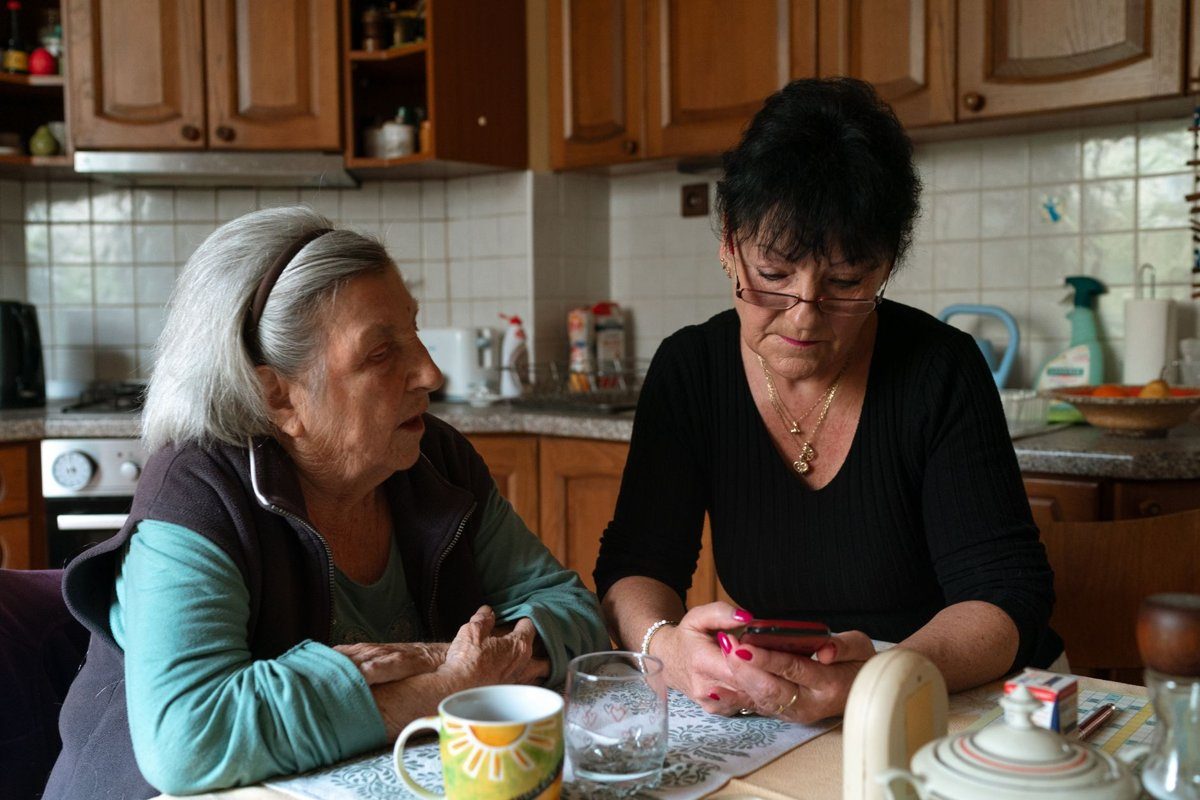 A caregiver in a black sweater checks her phone while sitting beside an older woman at a kitchen table with tea cups.