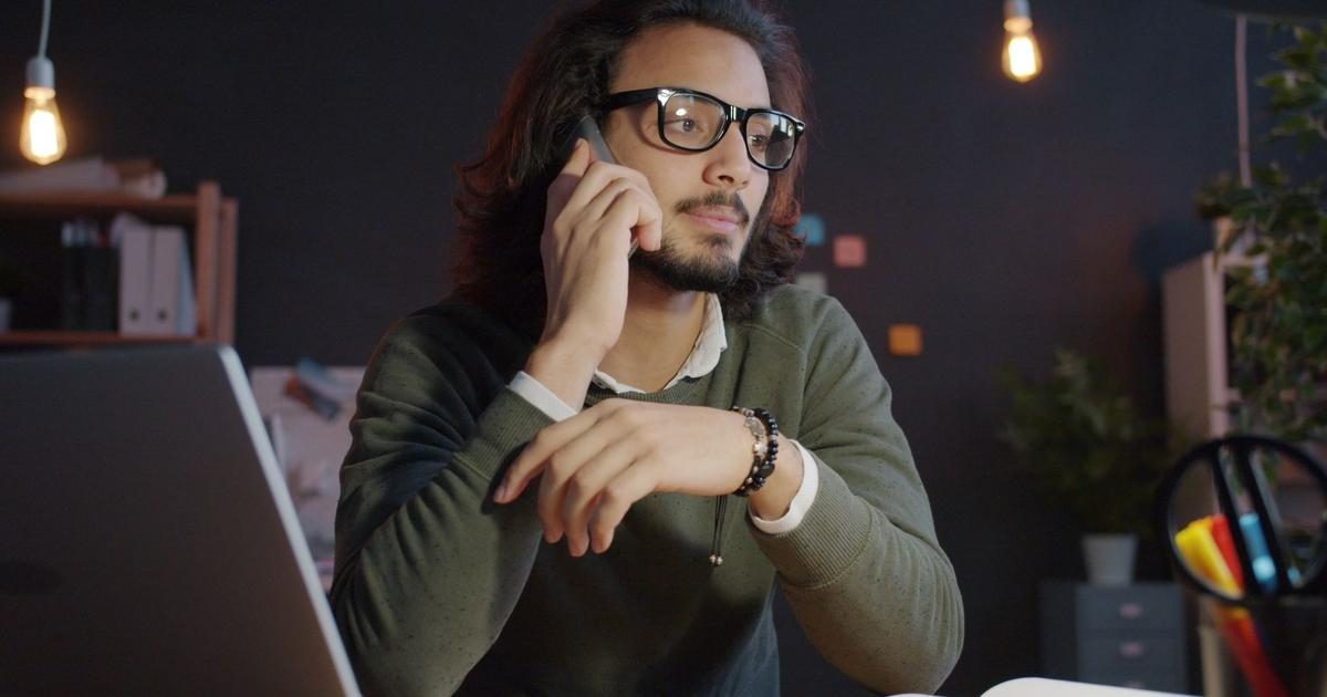 A man in glasses talks on his phone at a desk in a dim office lit by hanging bulbs late in the evening.
