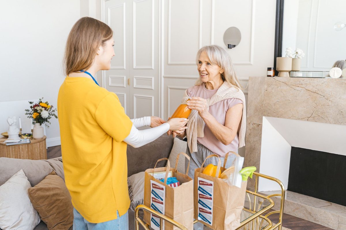 A home care aide in a yellow shirt hands a bottle of juice to a smiling older woman beside bags of groceries in her living room.