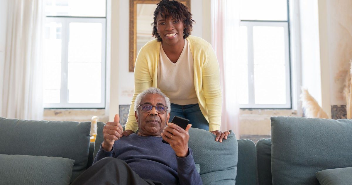 A smiling home care aide in a yellow cardigan stands behind an older man giving a thumbs up from his living room sofa.