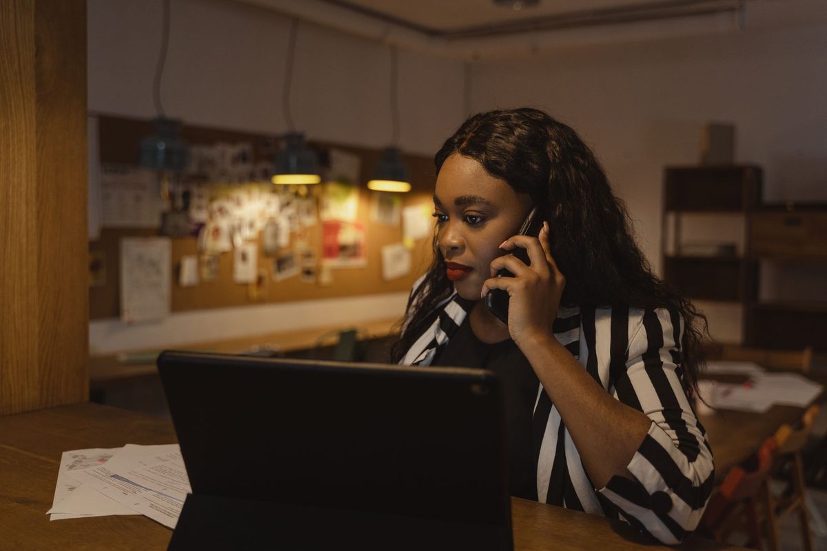 An office recruiter in a striped blazer talks on a desk phone while looking at her laptop in a small agency office.