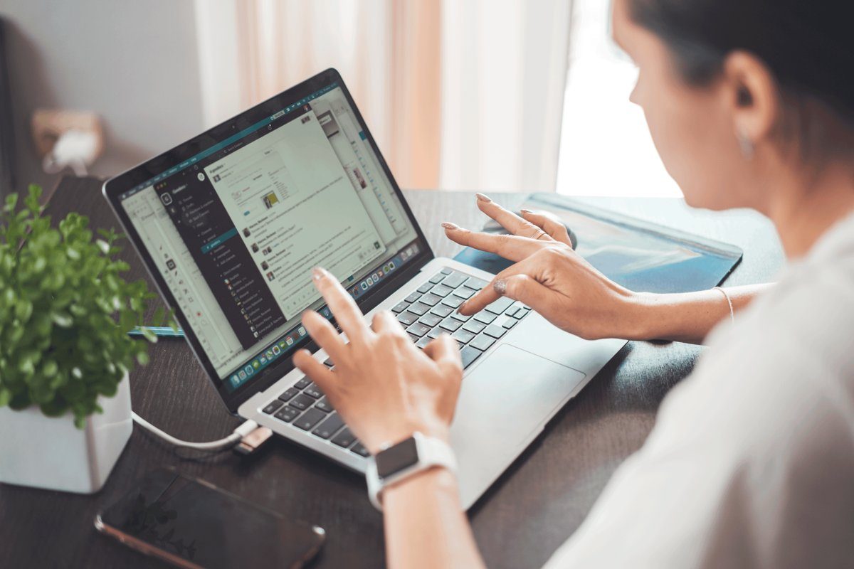 A woman types on a laptop at a table at home, with a phone and a small plant beside her.